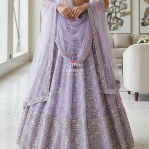K&ouml;nnte beinhalten: Ein lavendelfarbenes Lehenga-Choli-Set mit silbernen Pailletten. Rock und Crop-Top sind mit Stickereien verziert. Ein passendes, durchsichtiges Dupatta drapiert die Schultern. Der Text "CLOTHS STUDIOS" ist sichtbar.