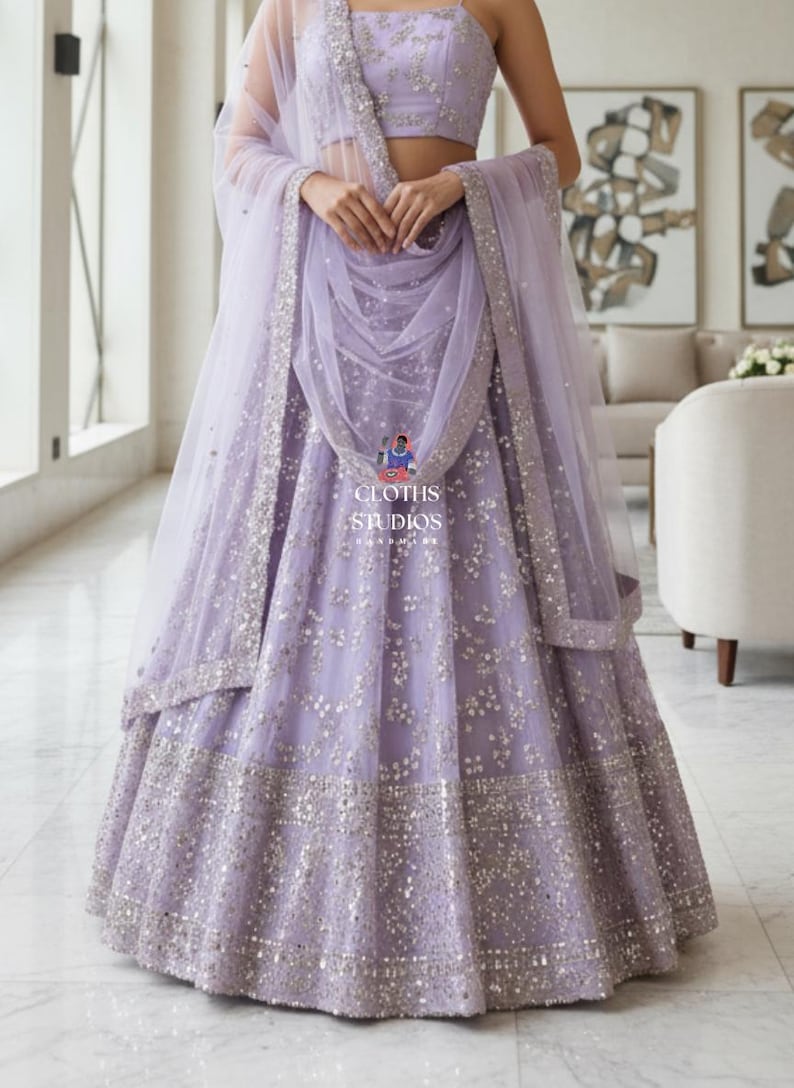 K&ouml;nnte beinhalten: Ein lavendelfarbenes Lehenga-Choli-Set mit silbernen Pailletten. Rock und Crop-Top sind mit Stickereien verziert. Ein passendes, durchsichtiges Dupatta drapiert die Schultern. Der Text "CLOTHS STUDIOS" ist sichtbar.