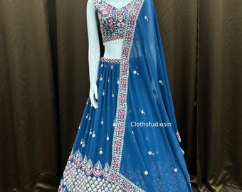 Puede incluir: Un conjunto lehenga choli azul turquesa sobre un maniquí. El conjunto incluye un top corto muy bordado, una falda a juego y un dupatta transparente. Patrones florales y geométricos en rosa, blanco y verde adornan los bordes. El texto "Clothstudiosin" es visible.