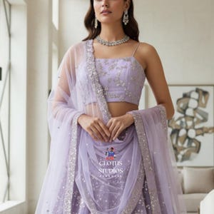 K&ouml;nnte beinhalten: Ein lavendelfarbenes formelles Kleid mit passendem transparentem Schal. Das Kleid hat aufwendige silberne Verzierungen und einen quadratischen Ausschnitt. Das Outfit wird mit einer silbernen Halskette und Ohrringen erg&auml;nzt. Der Text "CLOTHS STUDIOS HANDMADE" ist enthalten.