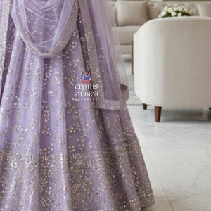 K&ouml;nnte beinhalten: Ein lavendelfarbenes formelles Kleid mit aufwendigen silbernen Verzierungen. Das Kleid hat einen weiten Rock und einen passenden, durchsichtigen Schal. Das Design umfasst ein detailliertes Muster aus kleinen, funkelnden Akzenten, die einen luxuri&ouml;sen Look erzeugen. Der Text "CLOTHS STUDIOS" ist enthalten.