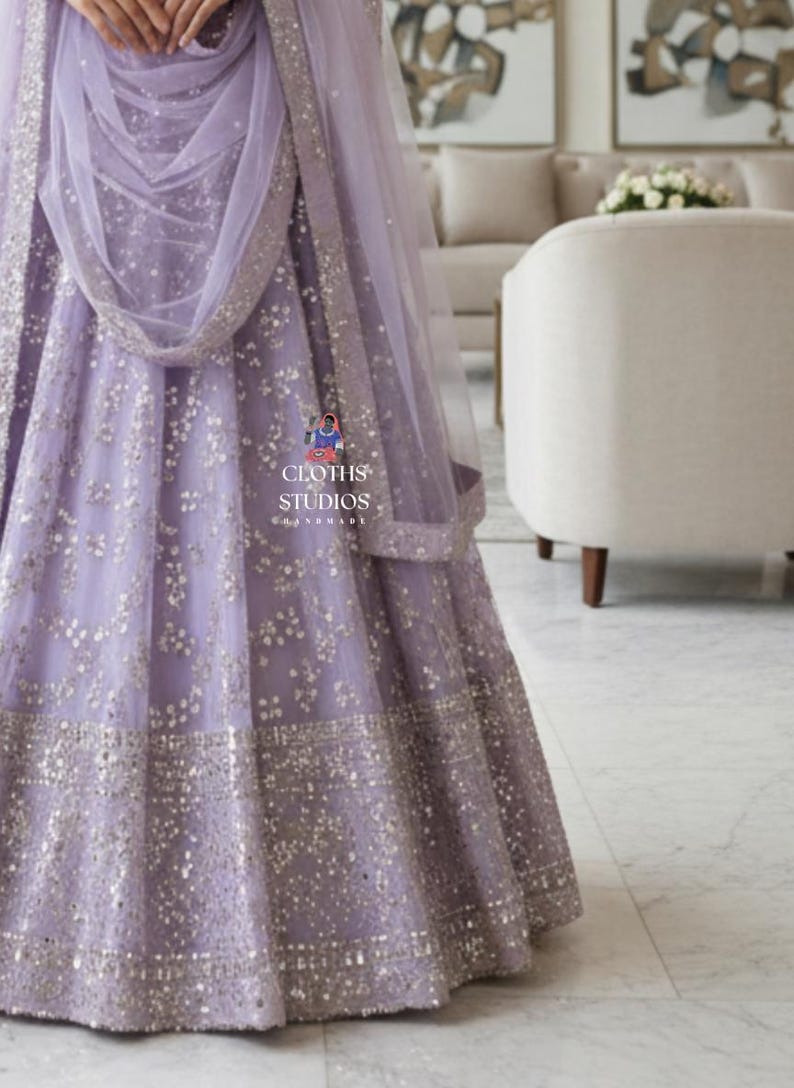 K&ouml;nnte beinhalten: Ein lavendelfarbenes formelles Kleid mit aufwendigen silbernen Verzierungen. Das Kleid hat einen weiten Rock und einen passenden, durchsichtigen Schal. Das Design umfasst ein detailliertes Muster aus kleinen, funkelnden Akzenten, die einen luxuri&ouml;sen Look erzeugen. Der Text "CLOTHS STUDIOS" ist enthalten.