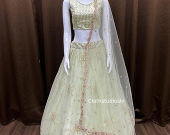 May include: A light green lehenga set on a mannequin. The set includes a sleeveless top, a long skirt, and a sheer dupatta. The outfit is embellished with gold-colored embroidery and a pink trim. The text "Clothstudiosin" is visible.