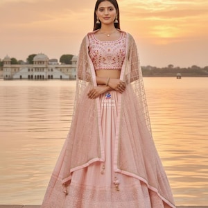 May include: A pale pink lehenga with detailed embroidery and a coordinating choli. The outfit includes a sheer, patterned dupatta. The background features a body of water and a building with a sunset in the background.