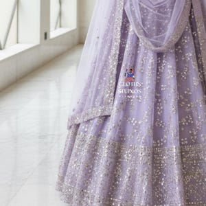 K&ouml;nnte beinhalten: Ein helllila Abendkleid mit silbernen Verzierungen. Das Kleid hat einen mehrschichtigen Rock und einen passenden, durchsichtigen Schal. Der Stoff ist mit verstreuten silbernen Pailletten verziert, und der Saum ist mit einem breiten Band aus silbernen Details versehen. Der Text "CLOTHS STUDIOS" ist enthalten.