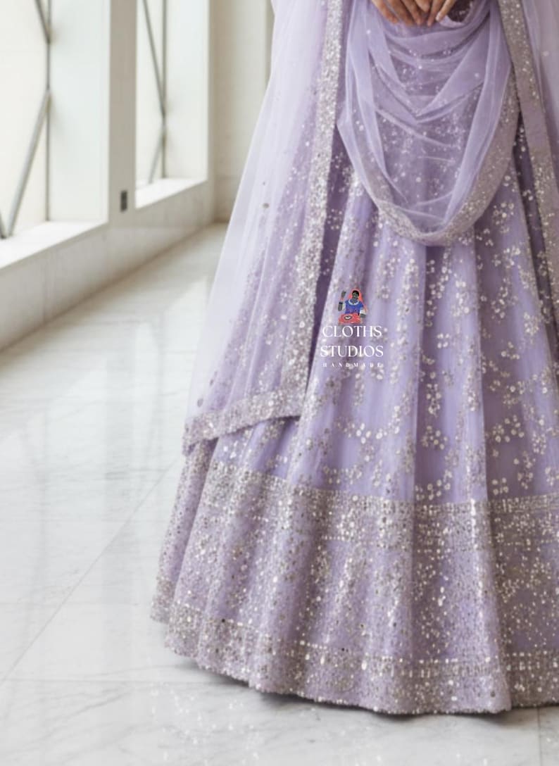 K&ouml;nnte beinhalten: Ein helllila Abendkleid mit silbernen Verzierungen. Das Kleid hat einen mehrschichtigen Rock und einen passenden, durchsichtigen Schal. Der Stoff ist mit verstreuten silbernen Pailletten verziert, und der Saum ist mit einem breiten Band aus silbernen Details versehen. Der Text "CLOTHS STUDIOS" ist enthalten.