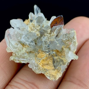 Puede incluir: Primer plano de un grupo de cristales de cuarzo transparente con una punta marrón rojiza. Los cristales están sobre una matriz beige y amarilla. Espécimen mineral natural.
