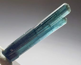 Puede incluir: Cristal de turmalina azul-verdosa de forma cilíndrica. La gema es translúcida, mostrando estrías internas y un sutil gradiente de color. El cristal se presenta sobre un fondo neutro, resaltando su belleza natural.