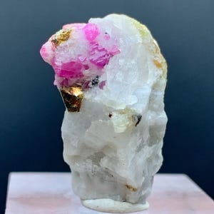 May include: A raw, uncut mineral specimen featuring a cluster of vibrant pink crystals embedded in a matrix of white and gold-colored stone. The rough texture and natural formation of the mineral are clearly visible, showcasing its unique geological characteristics.