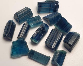 May include: A collection of raw, blue tourmaline crystals. The crystals vary in size and shape, with a deep blue color and visible striations. The gemstones are scattered on a white surface, showcasing their natural, unpolished state.