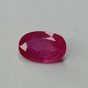 Peut inclure: Une pierre précieuse de rubis de coupe ovale. Le rubis est d'un rouge profond et vibrant avec des facettes visibles. La pierre est posée sur un fond uni gris clair.