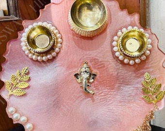 May include: A pink resin decorative tray with gold accents, featuring three gold bowls, faux pearls, and gold leaf designs. A small gold Ganesh statue is centered on the tray. The tray has a scalloped edge.