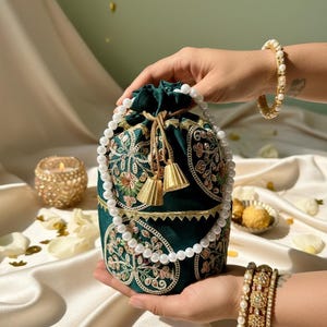 May include: An emerald green drawstring bag with intricate floral embroidery and gold tassels. A white pearl necklace and gold bracelets adorn the image. The bag is held in a person's hands, showcasing its design and craftsmanship.