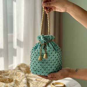 May include: A teal drawstring bag with a beaded handle and black embroidered details. The bag has a textured appearance and is held up by a beaded gold handle. Two small gold tassels hang from the drawstring closure.