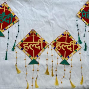 May include: Red and gold kite-shaped decorations with green and yellow details. The ornaments have text in a foreign language and are decorated with tassels and beaded strings, possibly for a cultural event.