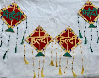 May include: Red and gold kite-shaped decorations with green and yellow details. The ornaments have text in a foreign language and are decorated with tassels and beaded strings, possibly for a cultural event.