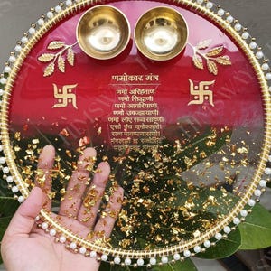 May include: A decorative red and gold tray with two small brass bowls. The tray features gold leaf accents, a beaded edge, and text in a foreign language. The design includes gold leaf and two symbols.