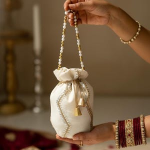 May include: A white drawstring pouch with gold and silver detailing and a beaded handle. The pouch is held by a person, highlighting its elegant design. Gold tassels hang from the drawstring closure, adding a touch of luxury. The pouch is likely used for carrying small items.