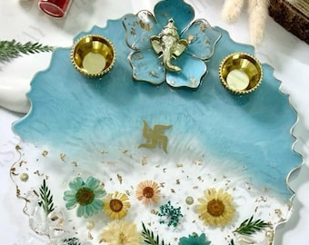 May include: A decorative resin tray with a light blue and white design, featuring a gold Ganesh statue and two small gold bowls. The tray is adorned with dried flowers and gold leaf accents, and includes two small glass jars.