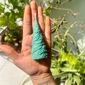 May include: A teal-colored, tree-shaped candle with a textured surface. The candle has a white wick and is held in a hand. The background features green foliage.