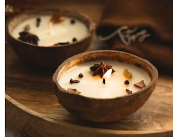 Può includere: Due candele accese in gusci di cocco. Le candele hanno una cera bianca cremosa, decorate con spezie e anice stellato. I gusci sono su un vassoio di legno. Sono un elemento di arredo naturale.