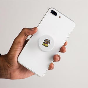 May include: A white smartphone with a round, white phone grip attached to the back. The phone grip has a cartoon graphic and the word "Single" in yellow. The phone is held in a hand.