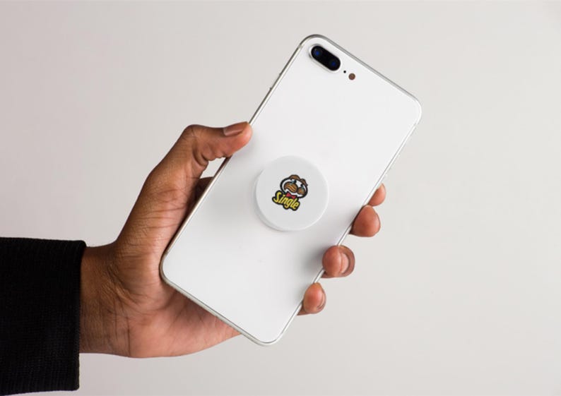 May include: A white smartphone with a round, white phone grip attached to the back. The phone grip has a cartoon graphic and the word "Single" in yellow. The phone is held in a hand.
