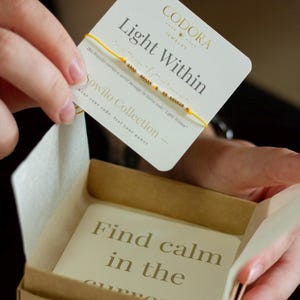 Puede incluir: Una pulsera de hilo amarillo con cuentas doradas se muestra con una tarjeta que dice "Light Within" y "Sowilo Collection". La pulsera está en una pequeña caja de cartón abierta con una tarjeta que dice "Find calm in the current".