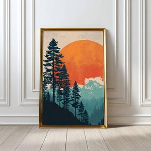 May include: Framed art print featuring a mountain landscape with a large orange sun setting behind silhouetted pine trees. The artwork has a gold frame and is displayed against a white wall and wooden floor.