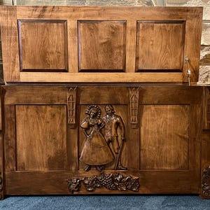 May include: A wooden chest with a hinged lid, featuring a carved relief of a couple and grapevines. The chest is stained a warm brown colour, with decorative carvings on the sides and front. The lid is divided into three panels.