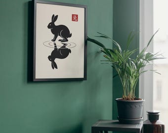Op de afbeelding: Een ingelijste kunstdruk met een zwart silhouet van een konijn dat boven zijn eigen reflectie in het water staat, tegen een lichtbeige achtergrond. Een klein rood vierkant met het witte Chinese karakter voor konijn staat rechtsboven.