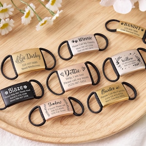 Op de afbeelding: Een verzameling gepersonaliseerde huisdier-ID-tags in verschillende kleuren, waaronder goud, zilver en zwart. Elke tag bevat een naam en contactgegevens, sommige met extra details. De tags liggen op een houten dienblad.