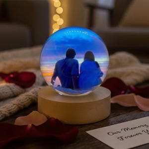 May include: A glowing glass sphere with a couple silhouette against a sunset scene, mounted on a wooden base. The sphere is surrounded by rose petals and a card that reads "Our Memory, Forever Yours."