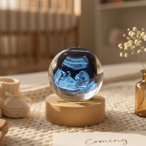 May include: A clear glass sphere displaying a baby's ultrasound image, resting on a wooden base. The sphere is illuminated, showcasing the baby's form. A card with the words "Coming Soon" is visible.
