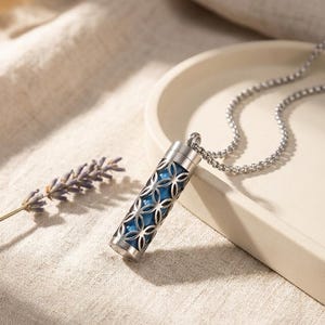 Puede incluir: Un collar con colgante plateado con un dije cilíndrico. El dije presenta un interior azul visible a través de un patrón calado decorativo. El collar se exhibe sobre una superficie de tono neutro con una ramita de lavanda.