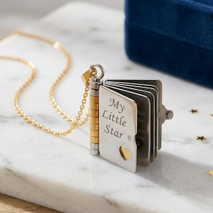 May include: A silver and gold locket necklace in the shape of a small book. The book has the words "My Little Star" engraved on the cover, with a heart cutout. The necklace chain is gold.
