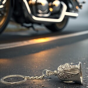 May include: A silver motorcycle bell keychain with an angel wing design lies on asphalt. A motorcycle is in the background, with chrome and gold accents. The bell is a common biker accessory.