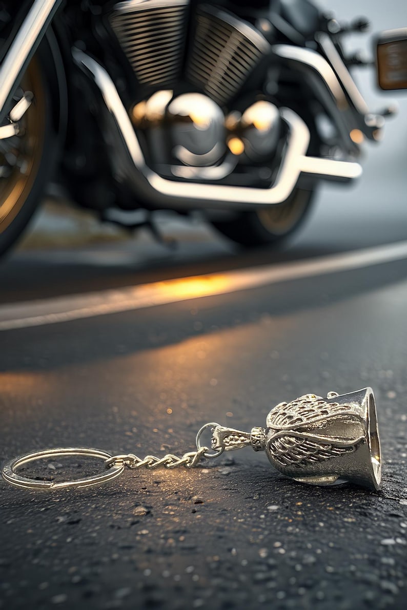 May include: A silver motorcycle bell keychain with an angel wing design lies on asphalt. A motorcycle is in the background, with chrome and gold accents. The bell is a common biker accessory.