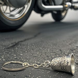 May include: A silver motorcycle bell keychain with the words "COME BACK SAFE TO ME" engraved on it, lying on a textured asphalt surface. A motorcycle wheel is visible in the background.