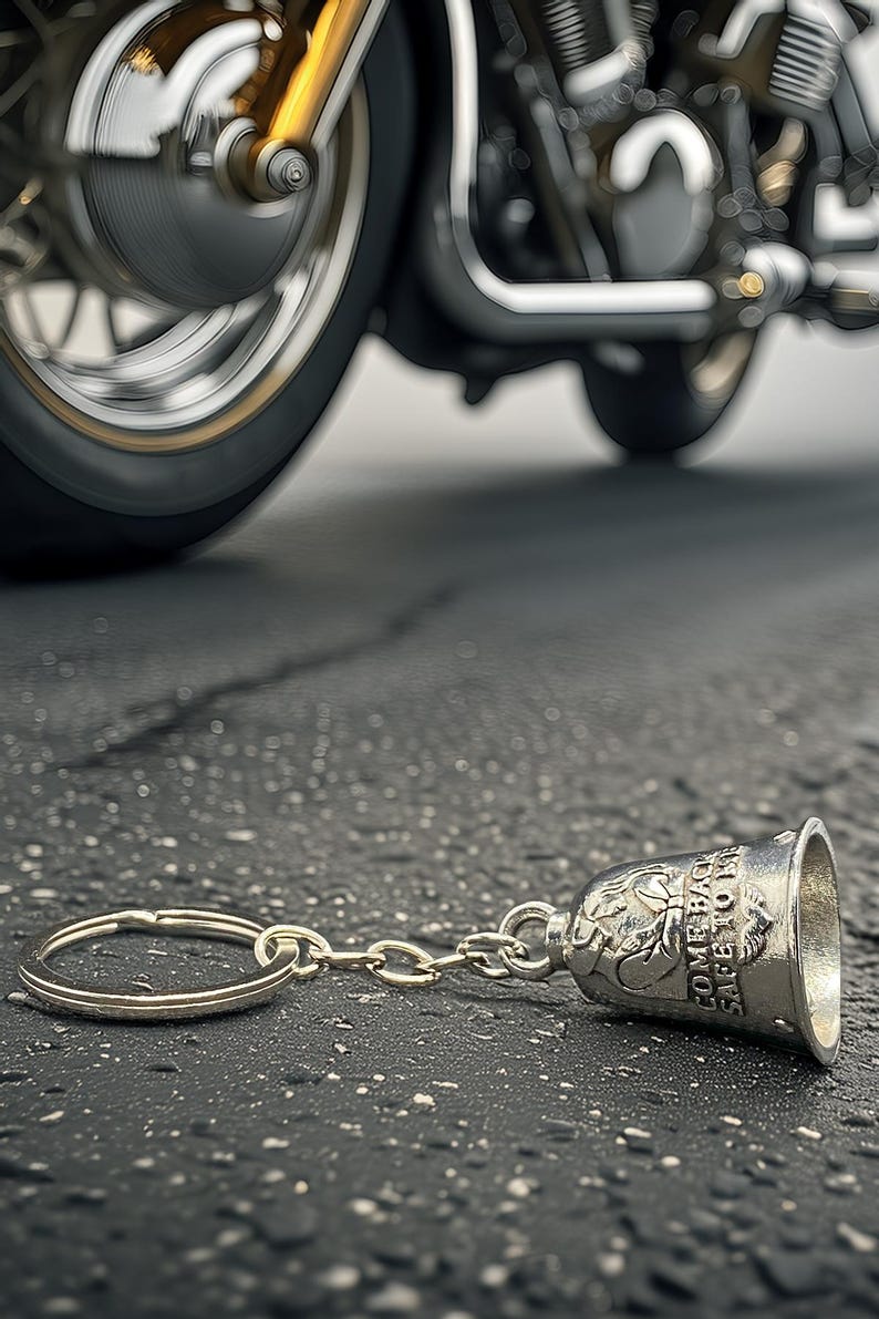 May include: A silver motorcycle bell keychain with the words "COME BACK SAFE TO ME" engraved on it, lying on a textured asphalt surface. A motorcycle wheel is visible in the background.