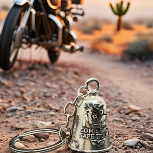 May include: A silver motorcycle bell keychain with the words "COME BACK SAFE TO" engraved on it. The bell is in focus, with a blurred motorcycle in the background. The scene is set in a desert landscape with a setting sun.