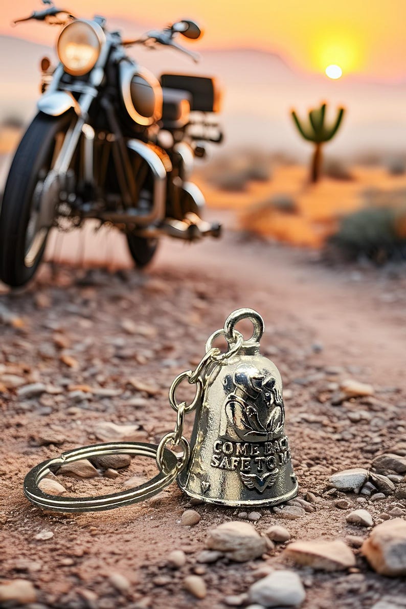 May include: A silver motorcycle bell keychain with the words "COME BACK SAFE TO" engraved on it. The bell is in focus, with a blurred motorcycle in the background. The scene is set in a desert landscape with a setting sun.