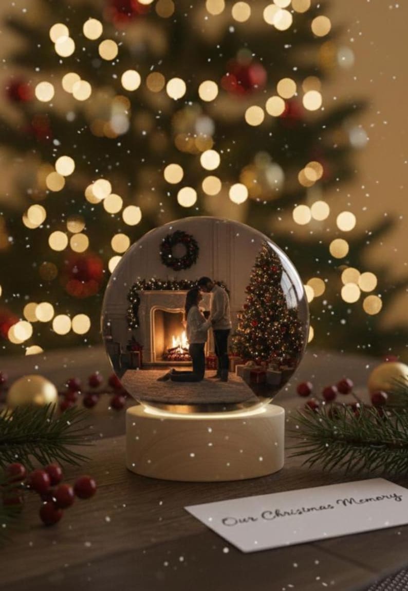 May include: A snow globe with a Christmas scene inside, featuring a couple in front of a fireplace and Christmas tree. The globe sits on a wooden base, with the text "Our Christmas Memory" on a card. Festive bokeh lights and decorations surround the globe.