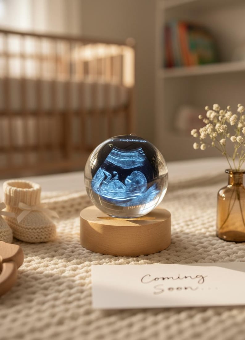 May include: A clear glass sphere displaying a baby's ultrasound image, resting on a wooden base. The sphere is illuminated, showcasing the baby's form. A card with the words "Coming Soon" is visible.