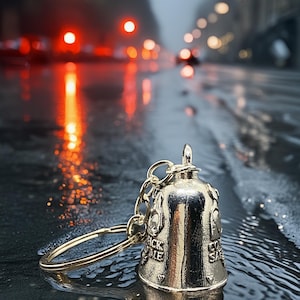 May include: A gold-colored bell keychain with the words "Ride Safe" and "Back Me" engraved on it. The bell is on a chain and ring. The background is a wet street with blurred lights.