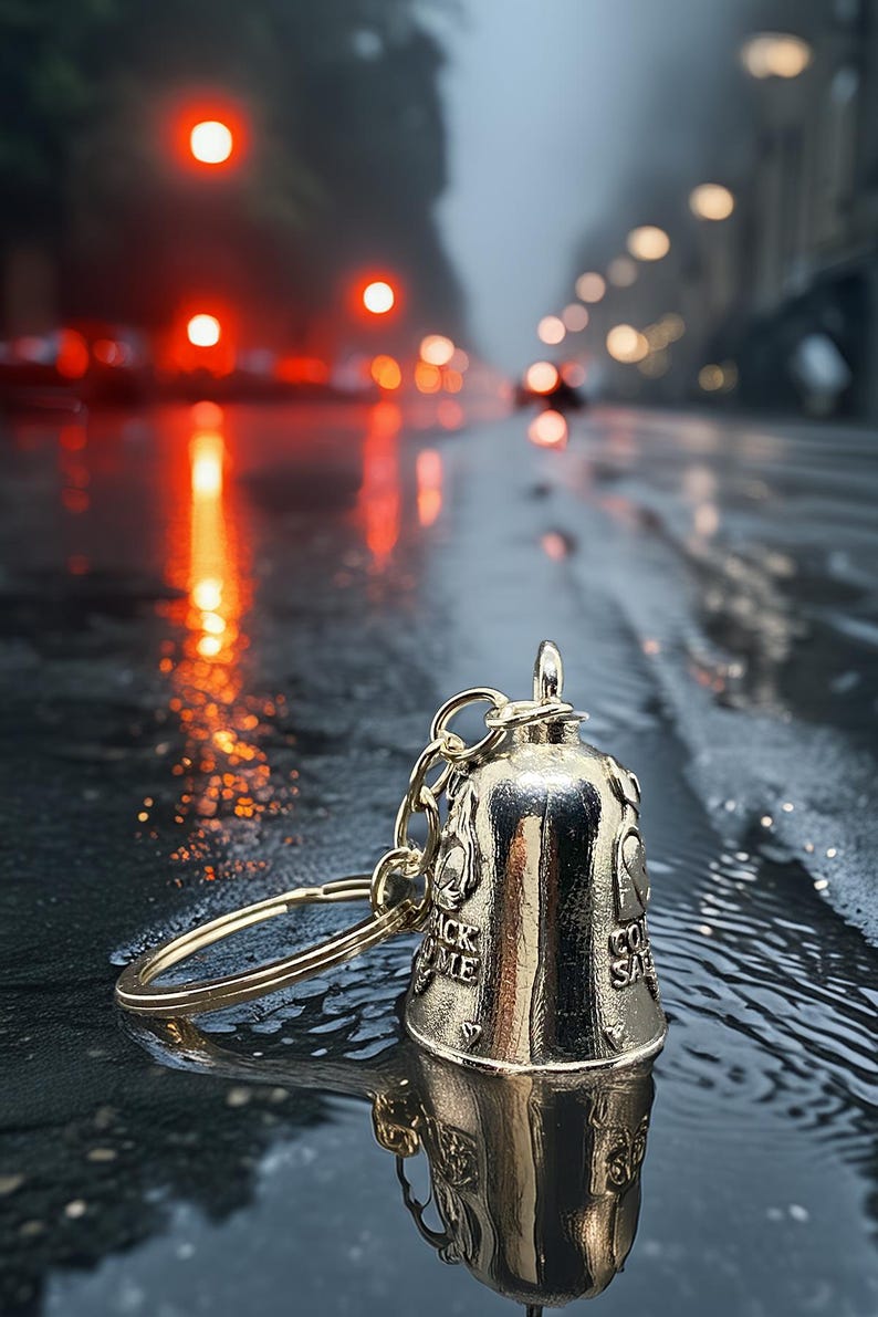May include: A gold-colored bell keychain with the words "Ride Safe" and "Back Me" engraved on it. The bell is on a chain and ring. The background is a wet street with blurred lights.