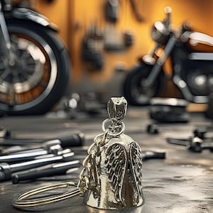 May include: A silver motorcycle bell with an angel wing design, hanging from a chain and ring. The bell is in focus, with a blurred background of a workshop and motorcycles. Tools are scattered on the work surface.