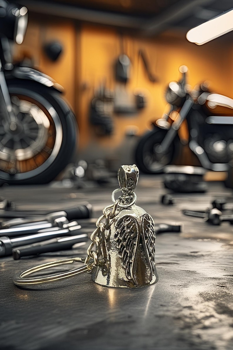 May include: A silver motorcycle bell with an angel wing design, hanging from a chain and ring. The bell is in focus, with a blurred background of a workshop and motorcycles. Tools are scattered on the work surface.