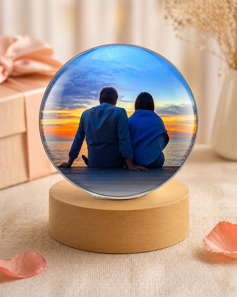 May include: A round, illuminated photo display featuring a couple watching a sunset over the water. The image is encased in a clear glass sphere, set on a wooden base. The background includes a gift box and dried flowers.