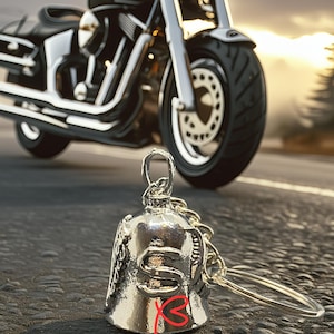May include: A silver motorcycle bell with a keychain, with the word "Safety" in the image. A black and chrome motorcycle is in the background. The bell is a common accessory for motorcycle riders.